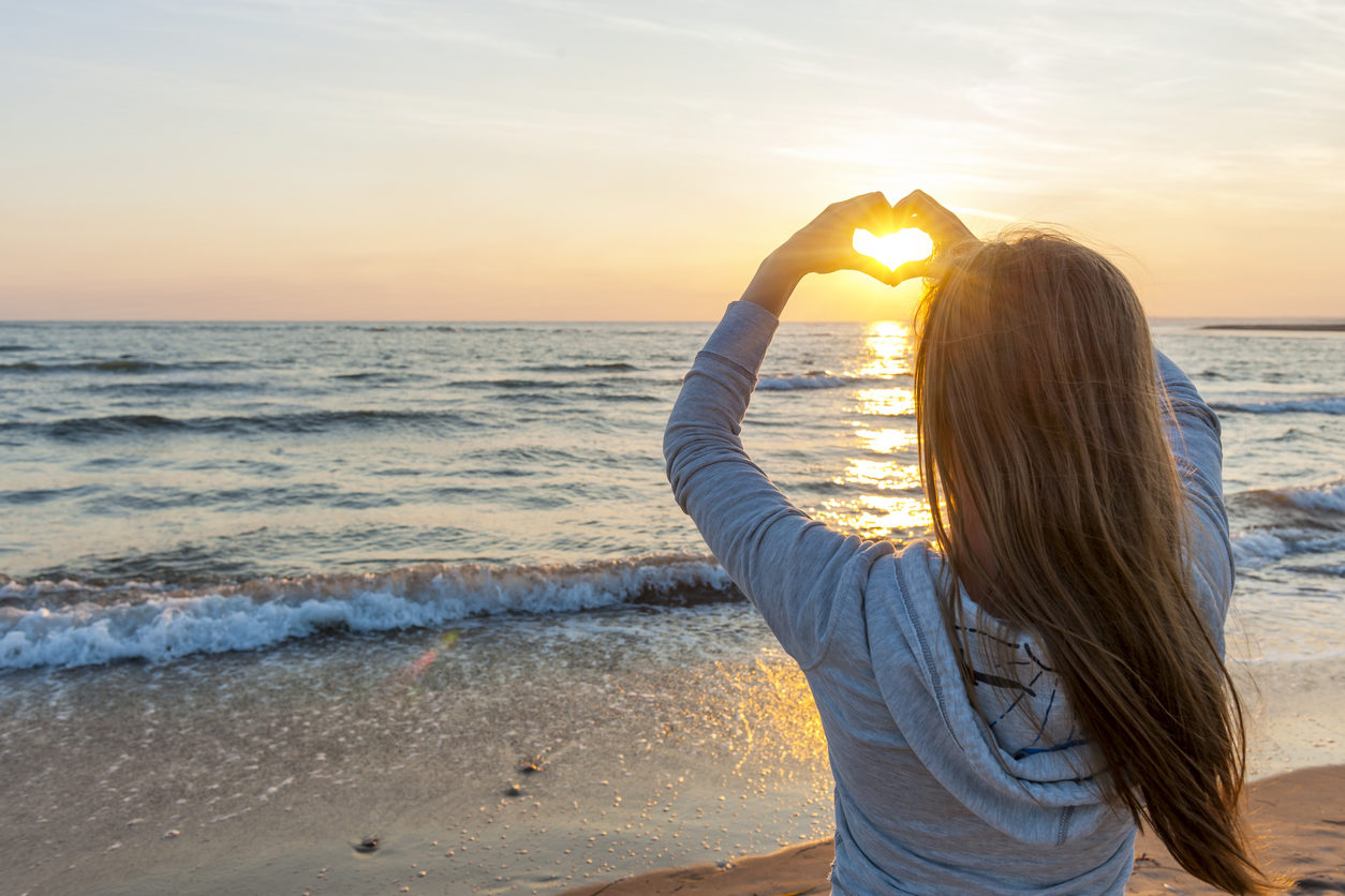 Woman making a heart shape with her hands against a beach sunset